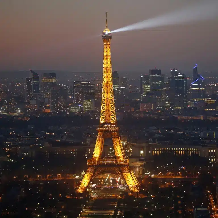 Tour Eiffel illuminée la nuit, avec son faisceau lumineux, surplombant la ville de Paris éclairée en arrière-plan.